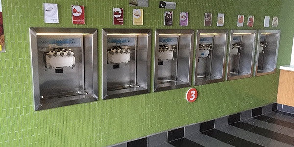 ice cream shop has green wall with 6 taylor ice cream machines inside the wall with colorful tag descriptions on top so customers know what flavor ice cream they are pouring into their cup.